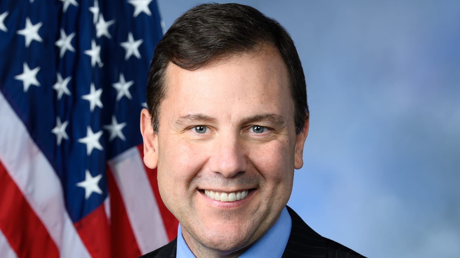 Tom Kean Jr. in a dark suit and red tie, smiling, in front of an American flag.
