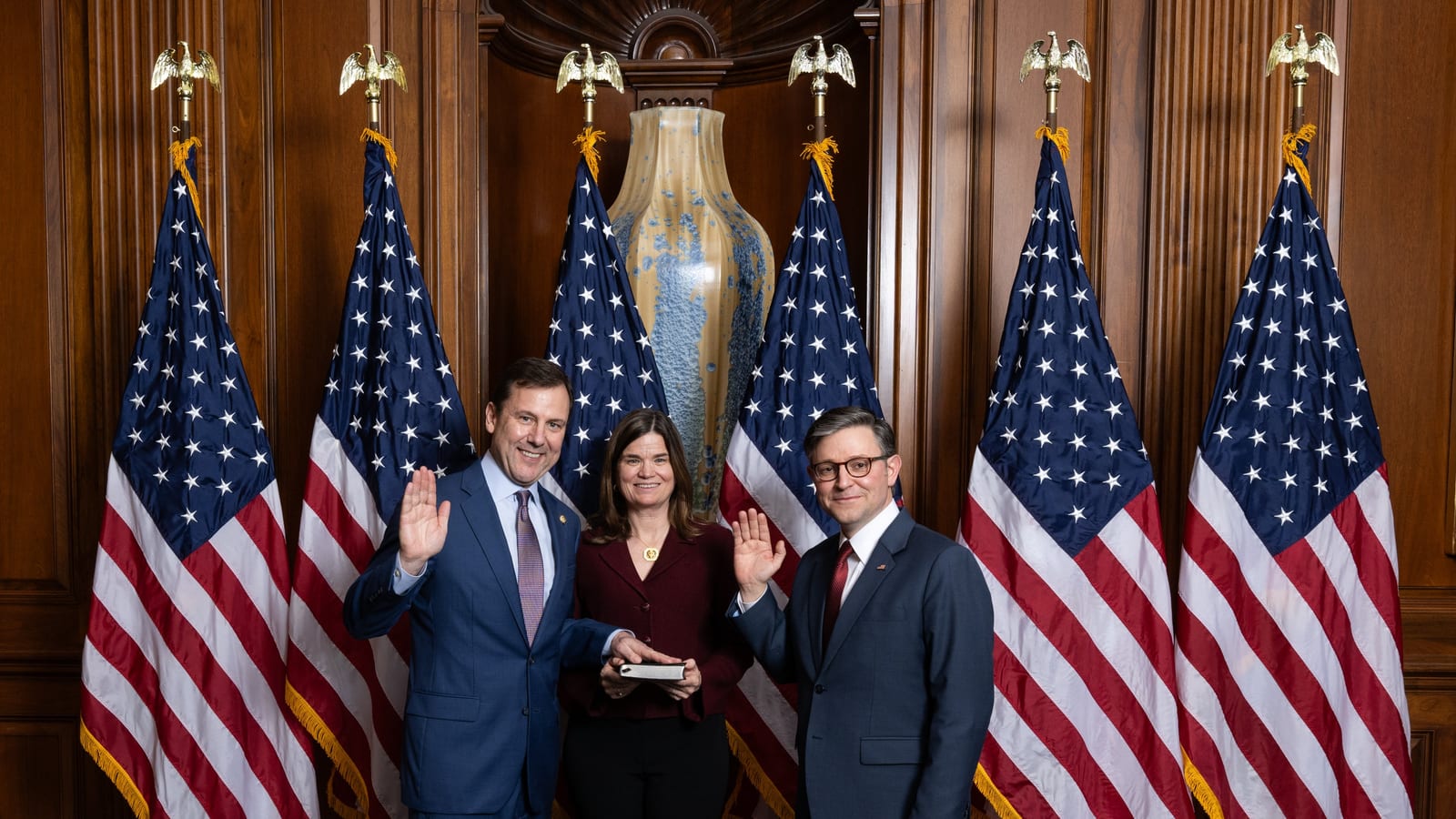 Tom Kean Jr. raises his right hand beside two other people in front of a row of American flags in a wood-panelled room.