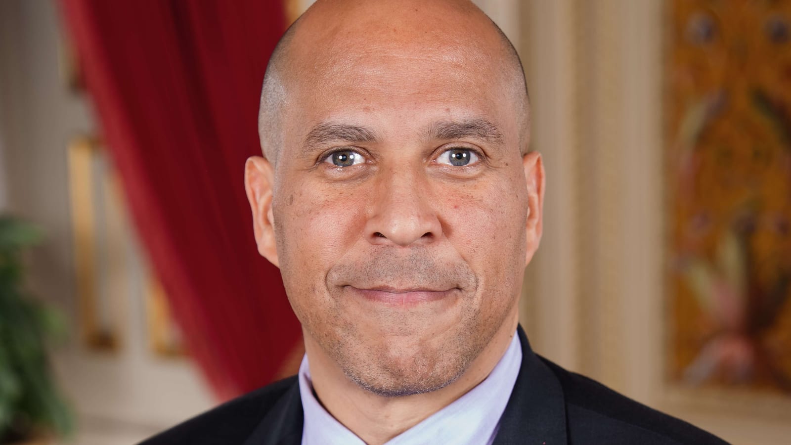 Cory Booker in a dark suit, light blue shirt and purple tie, smiling slightly in front of a red curtain in his official Senate portrait.