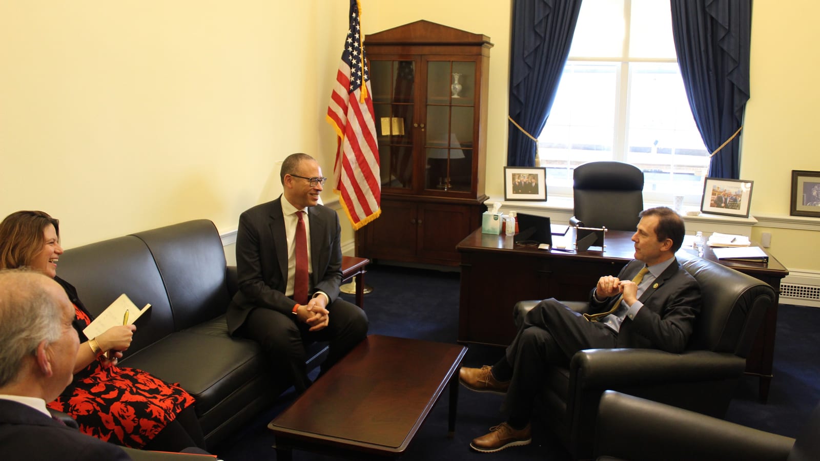 Four people talk in a congressional office with an American flag, a wooden cabinet and a window hung with blue drapes; a man in a dark suit sits in a leather armchair at right.