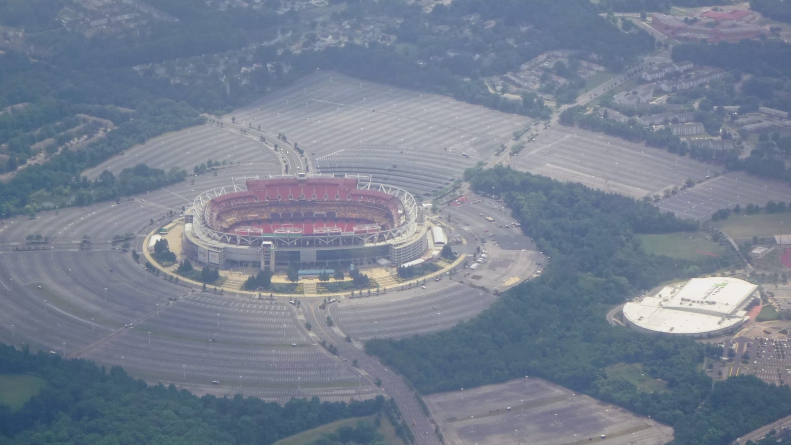 An aerial view of a large oval stadium with red seats in Landover, Maryland, ringed by empty parking lots and trees.