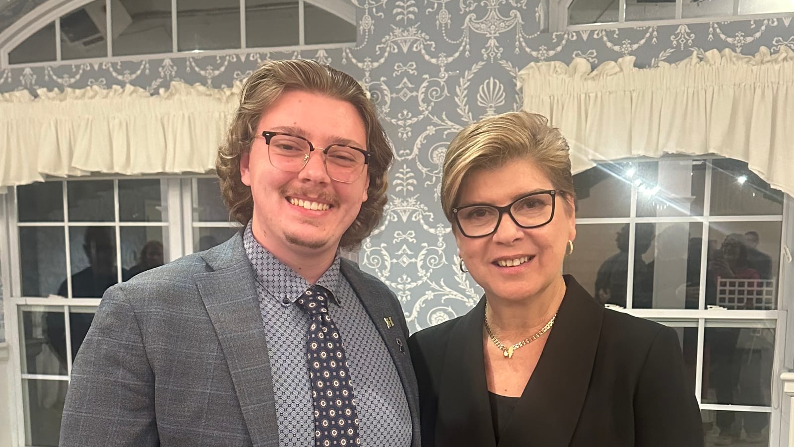 Nellie Pou, in a black jacket and glasses, stands beside Paul Duerr in front of patterned wallpaper and arched windows at an indoor campaign event.
