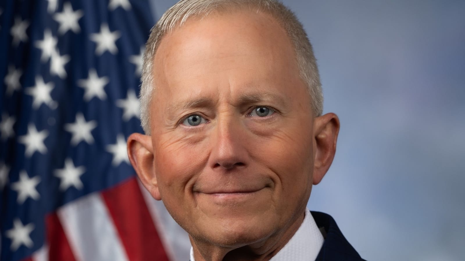 Jeff Van Drew in a navy suit, white shirt and yellow tie, smiling in front of an American flag and a blue backdrop.