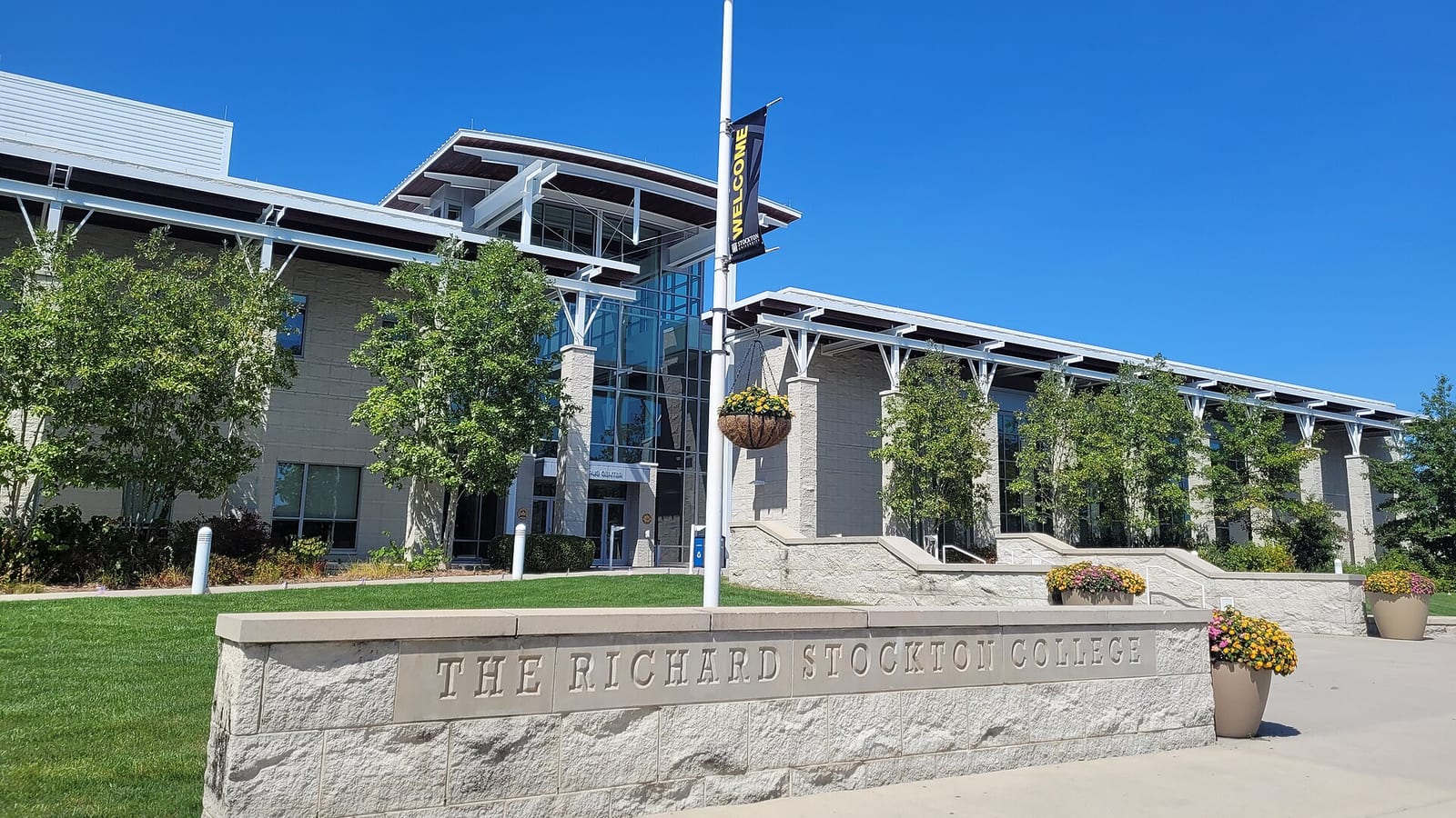 A low stone wall carved with the words The Richard Stockton College in front of a modern building with a glass entrance tower, a lamppost with a Welcome banner and trees under a clear blue sky.