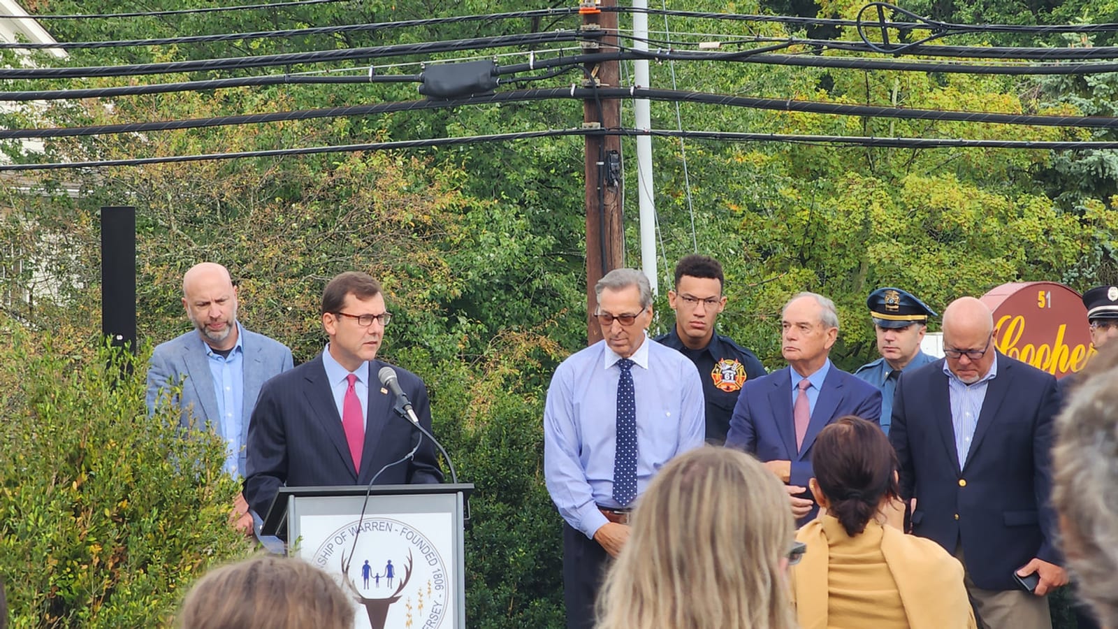 Tom Kean Jr. in a dark suit and red tie speaks at a lectern bearing the Township of Warren seal, with uniformed police and firefighters and other officials standing behind him.